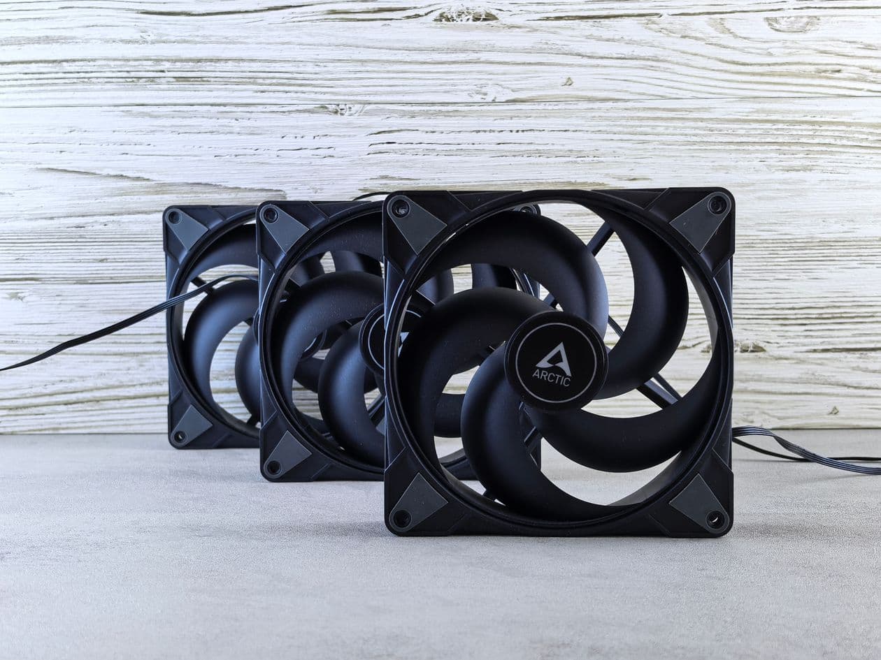 Three ARCTIC 120mm case fans laid out on a wooden surface, showing the blade profile and frame detail of a typical high-airflow gaming PC fan